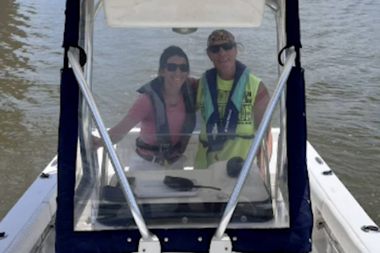Two people smiling on a boat, wearing life jackets and sunglasses, with water visible in the background.