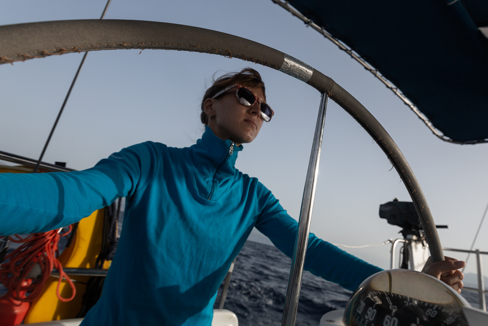 Woman in turquoise long-sleeve shirt and sunglasses steering a sailboat.