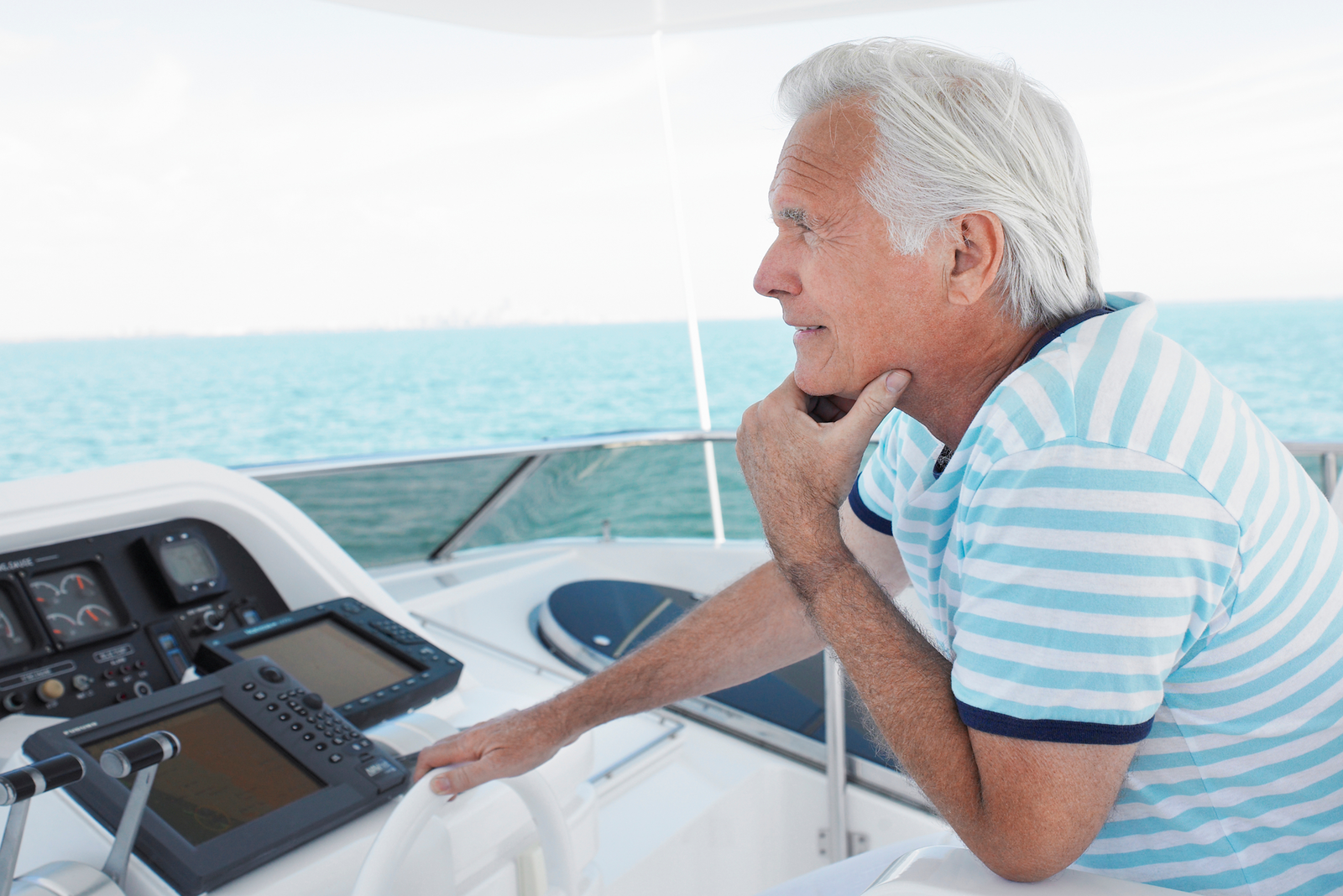 An older man with white hair wearing a blue and white striped shirt pilots a boat on open water.
