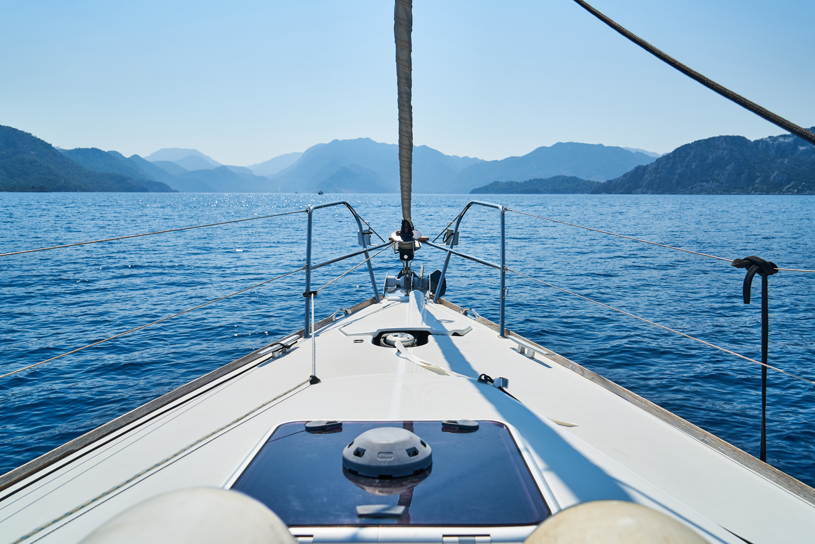 Front deck of a boat cruising on a calm blue sea, surrounded by distant mountains under a clear sunny sky.