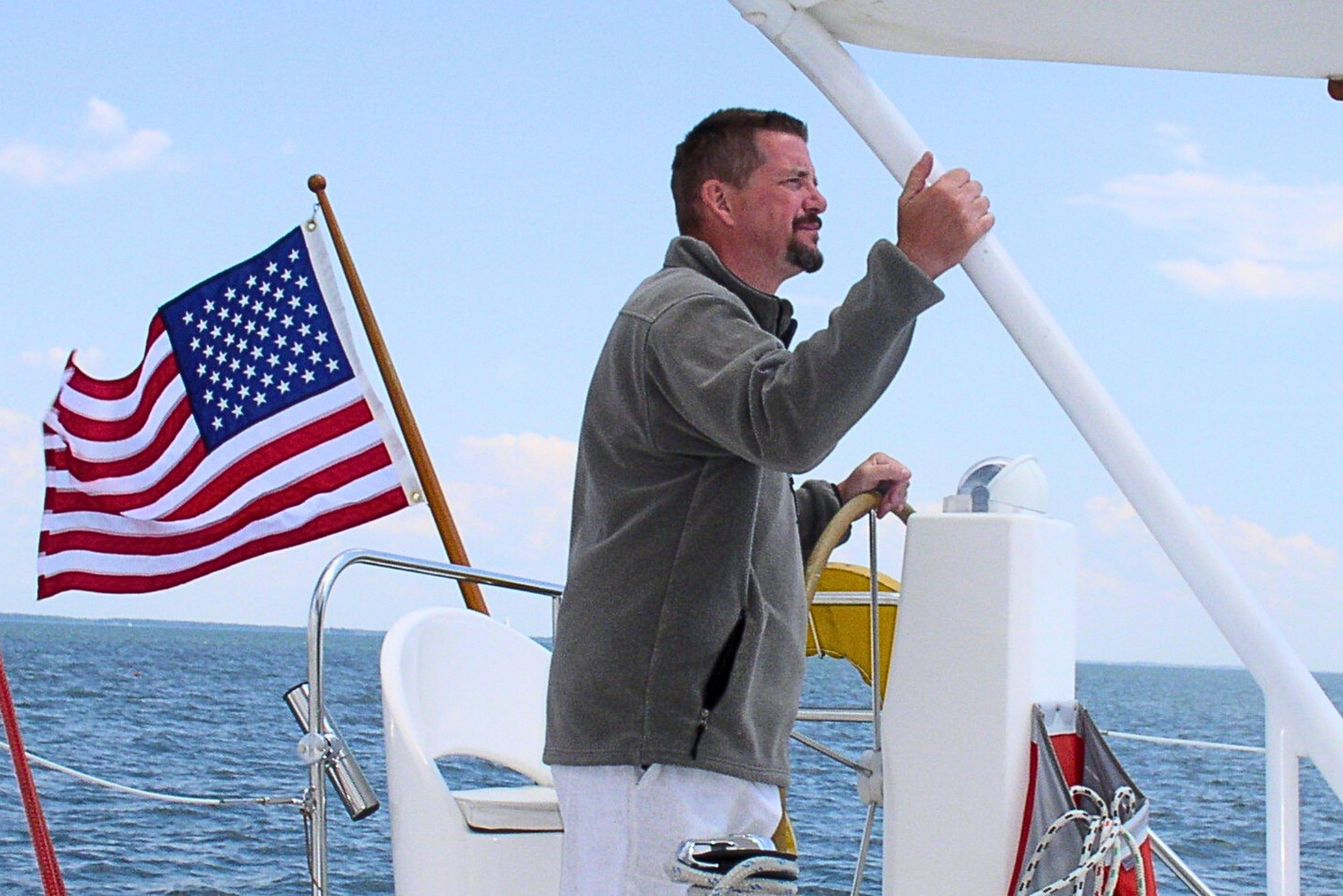Captain Bob Figular stands at the wheel of his boat, looking at the horizon, with an American flag waving in the wind behind him.
