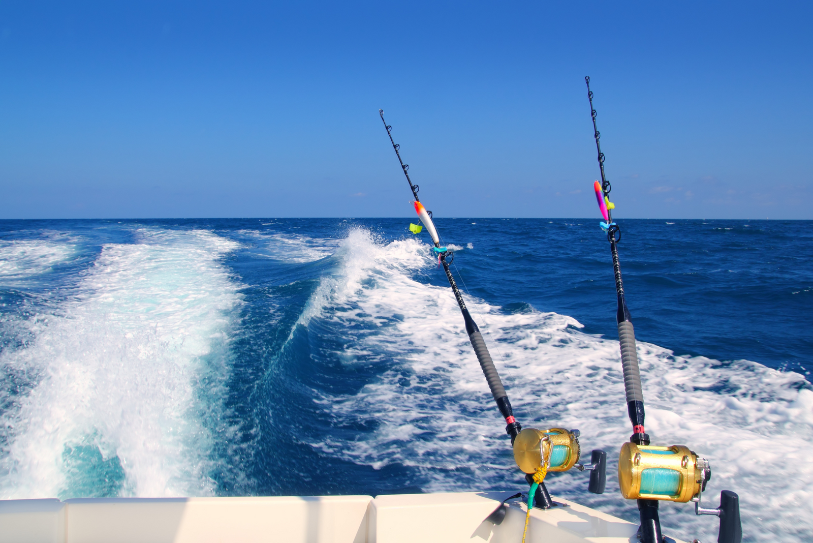Two fishing rods with reels mounted on a boat, leaving a trail of white foam on the deep blue ocean under a clear sky.