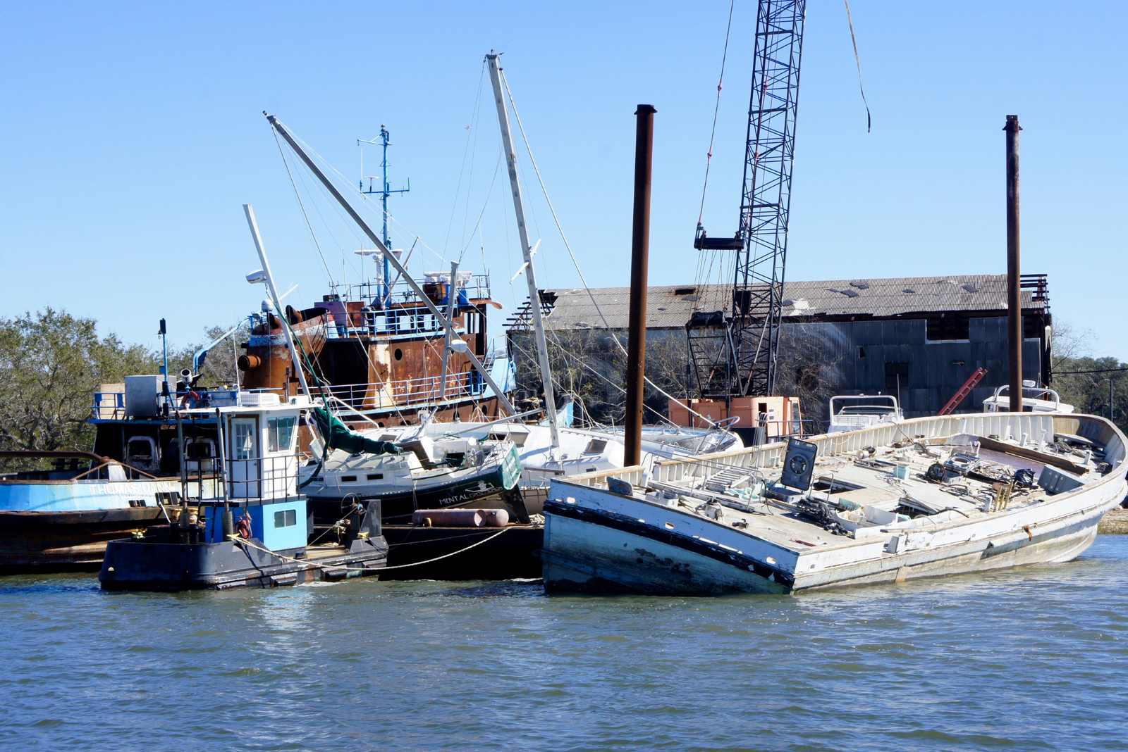 Cluster of damaged boats, including rusted and partially sunken vessels, near a dock with a crane.