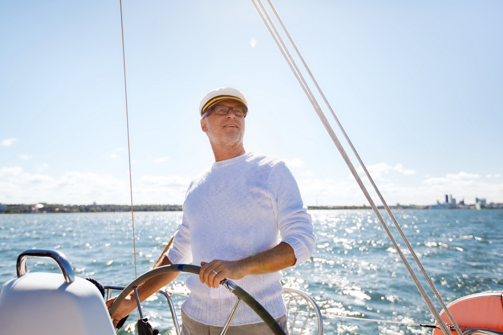 Captain in white sweater and sailing cap steering sailboat wheel on sunny day.