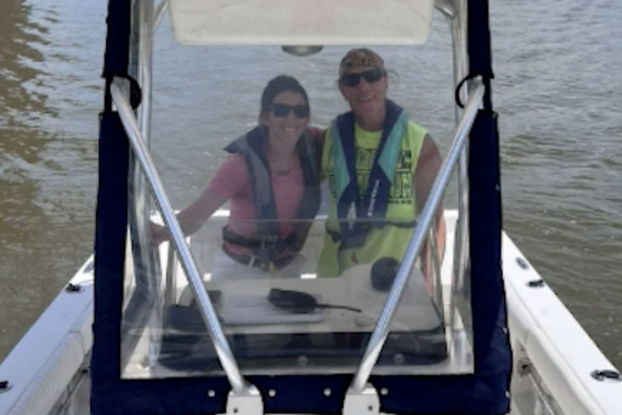 Two people smiling on a boat, wearing life jackets and sunglasses, with water visible in the background.