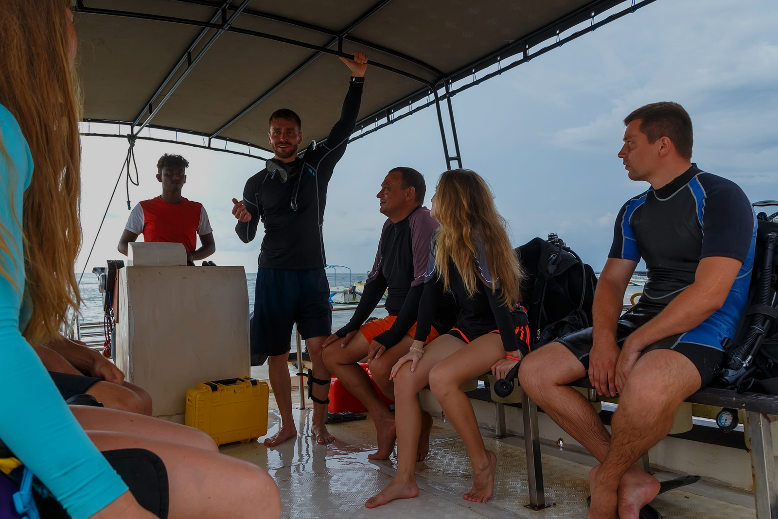 An instructor teaches a scuba diving lesson to attentive students on a boat deck, with the ocean visible behind them.