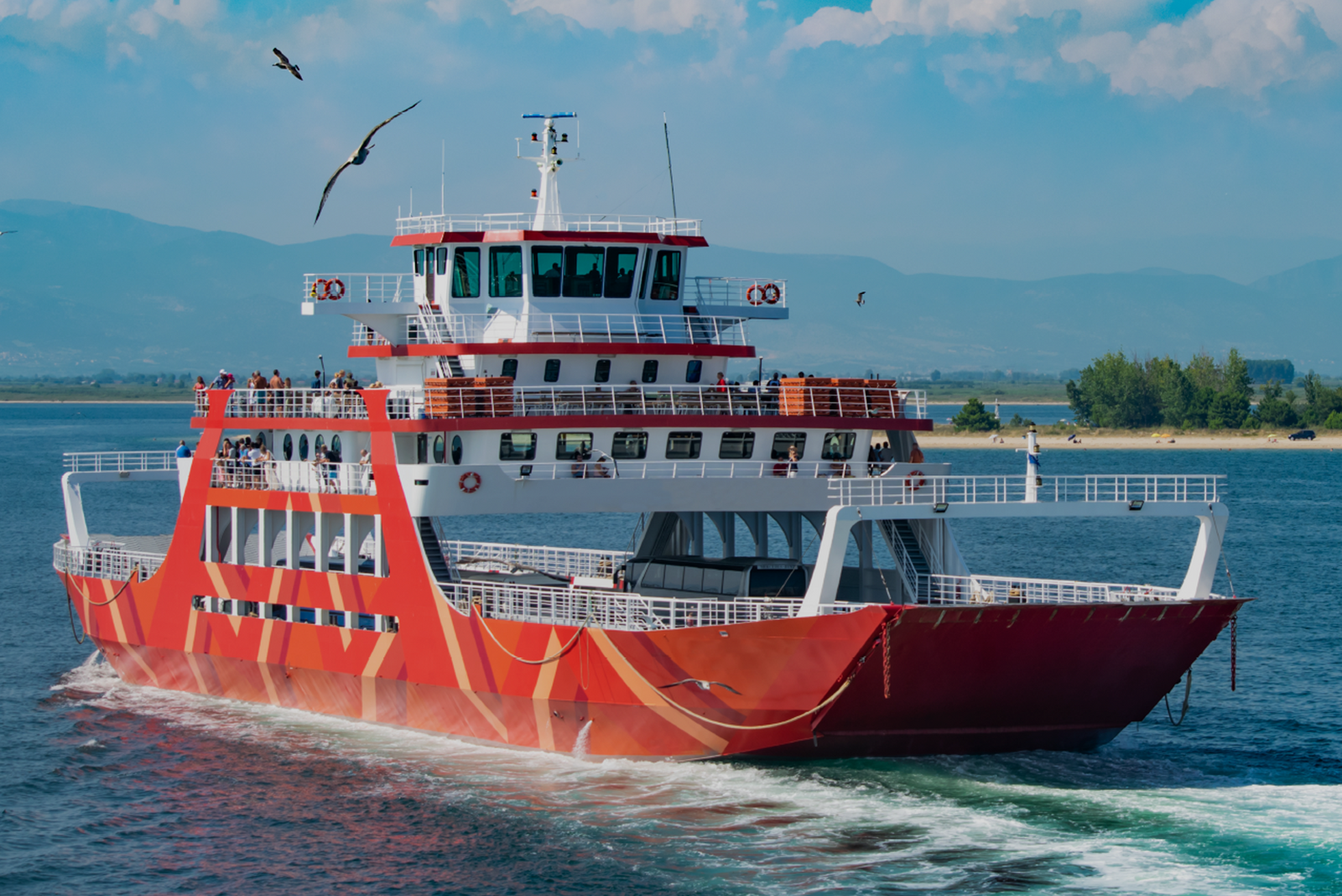 Red and white inspected vessel underway with passengers on upper decks and mountains in the distance.