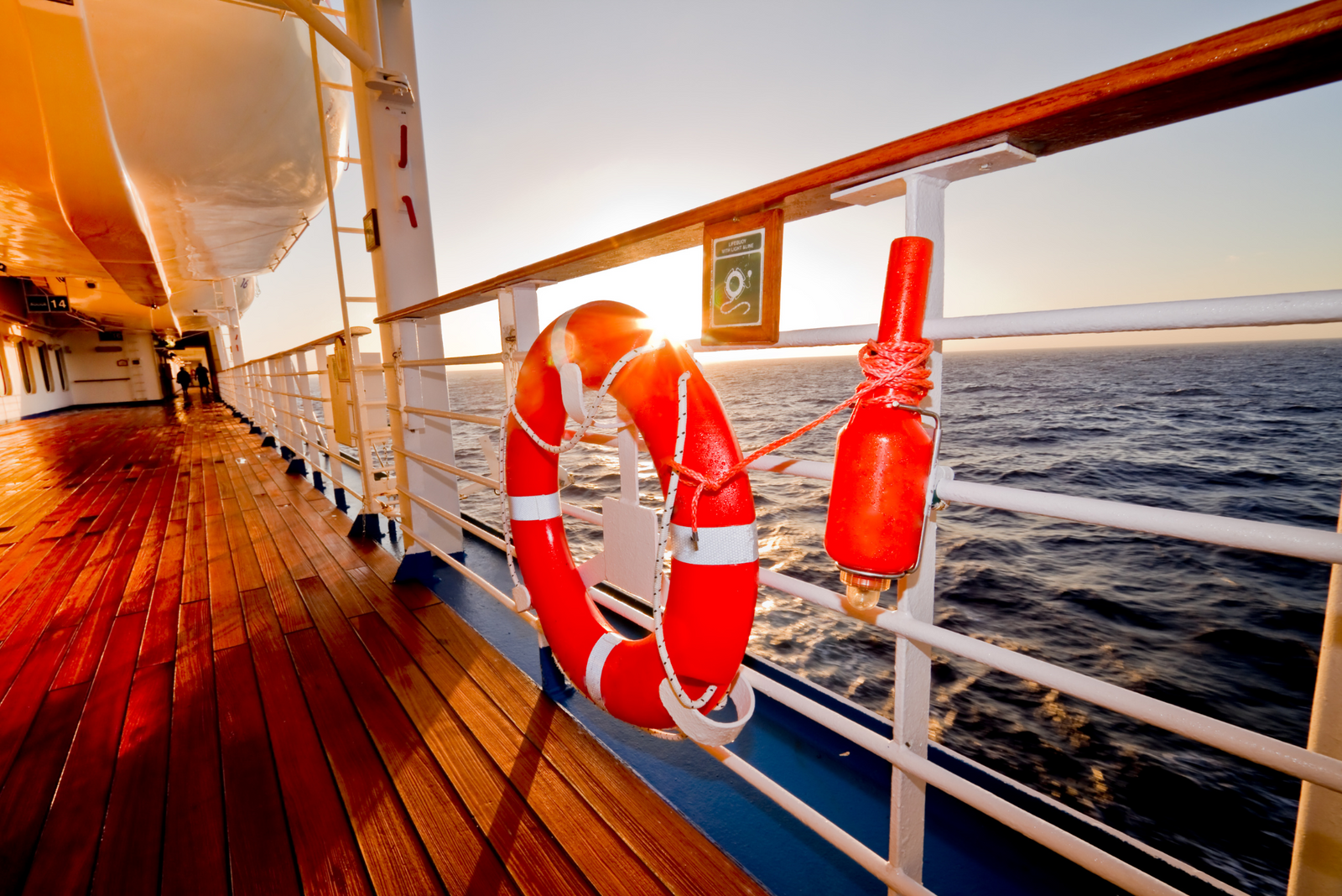 Life preserver and safety equipment on cruise ship deck at sunset, wooden deck and ocean view visible.