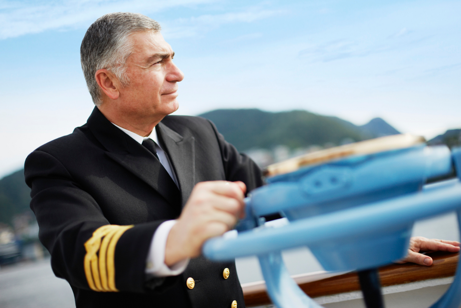 A boat captain in formal uniform with gold stripes looking ahead, standing at ship’s deck.