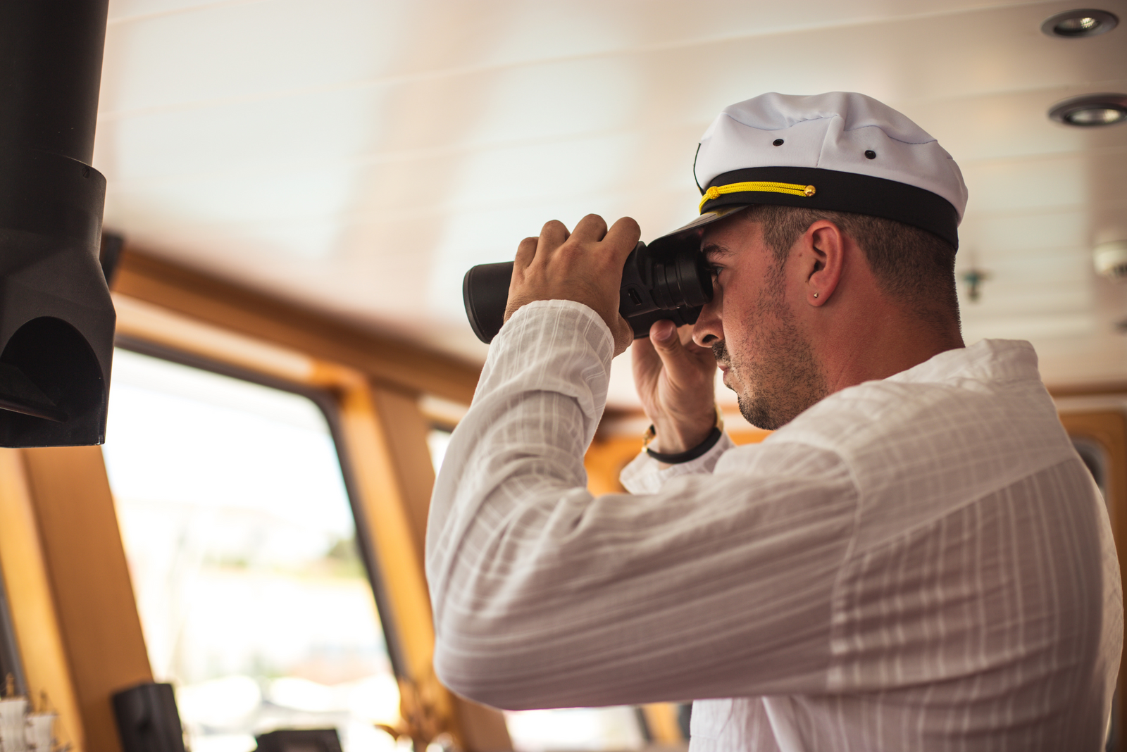 Ship captain in white uniform and cap using binoculars to scan horizon from inside vessel’s bridge cabin.