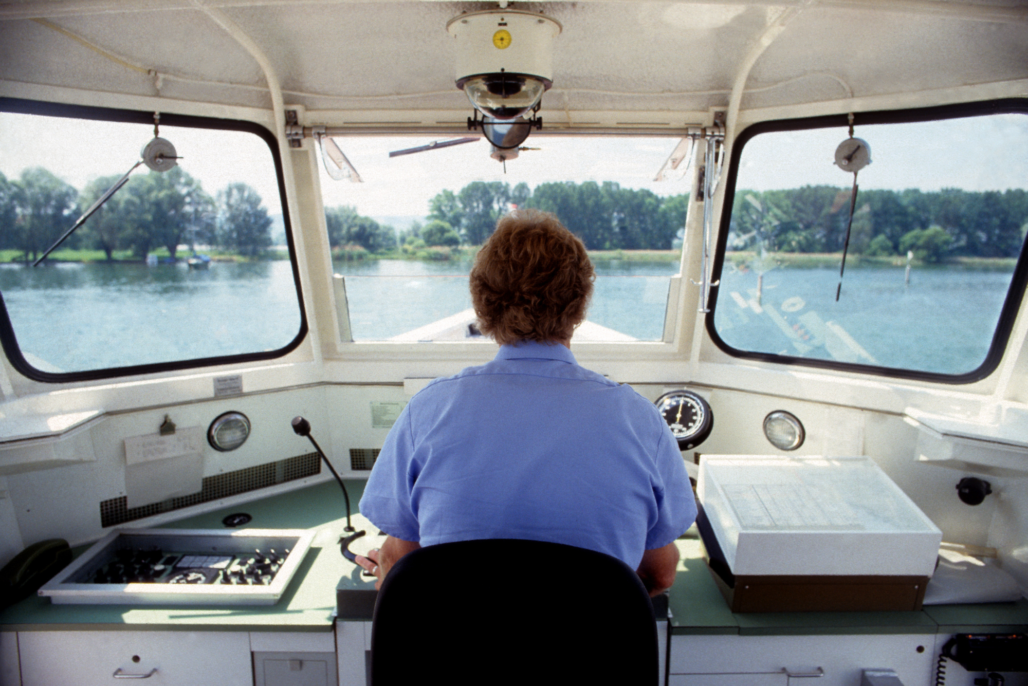 Boat captain wearing a blue shirt operating a vessel, viewed from behind in the control room.