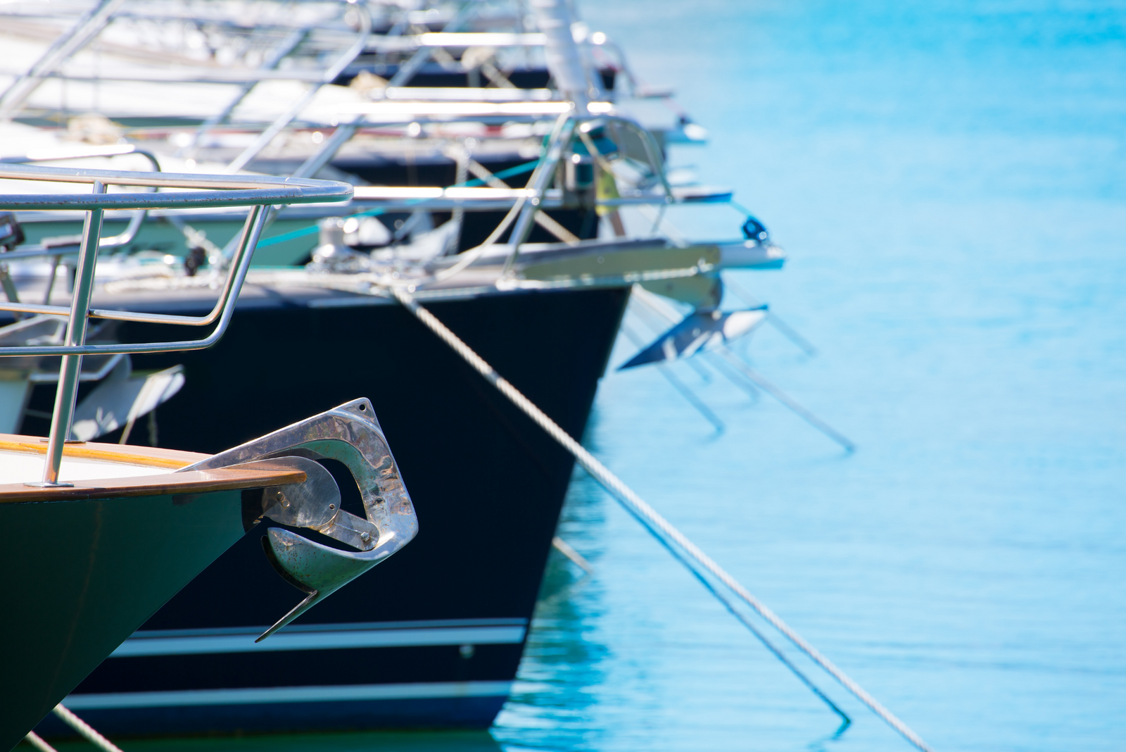 Line of anchored boats in marina, bow anchors visible, with boats moored diagonally against calm blue water.