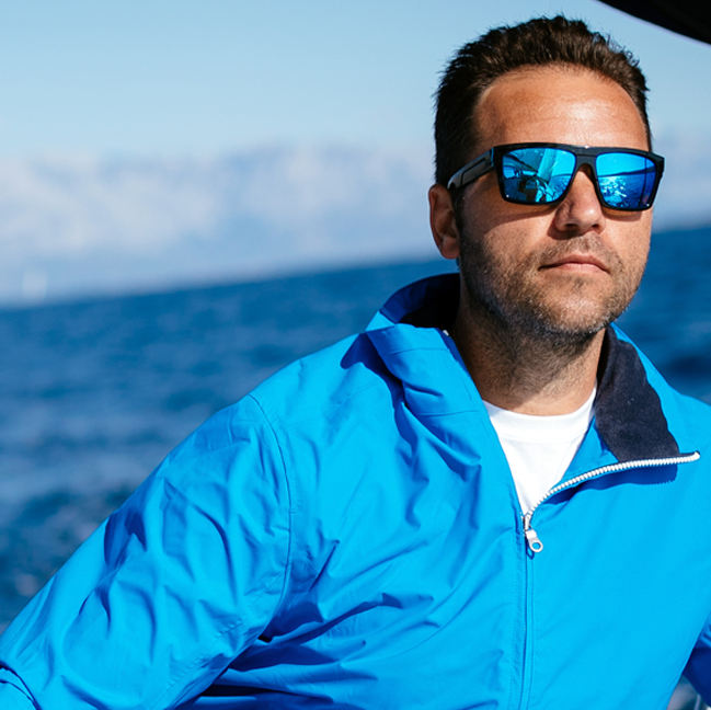 Man in a blue jacket and sunglasses on a boat with mountains in the background